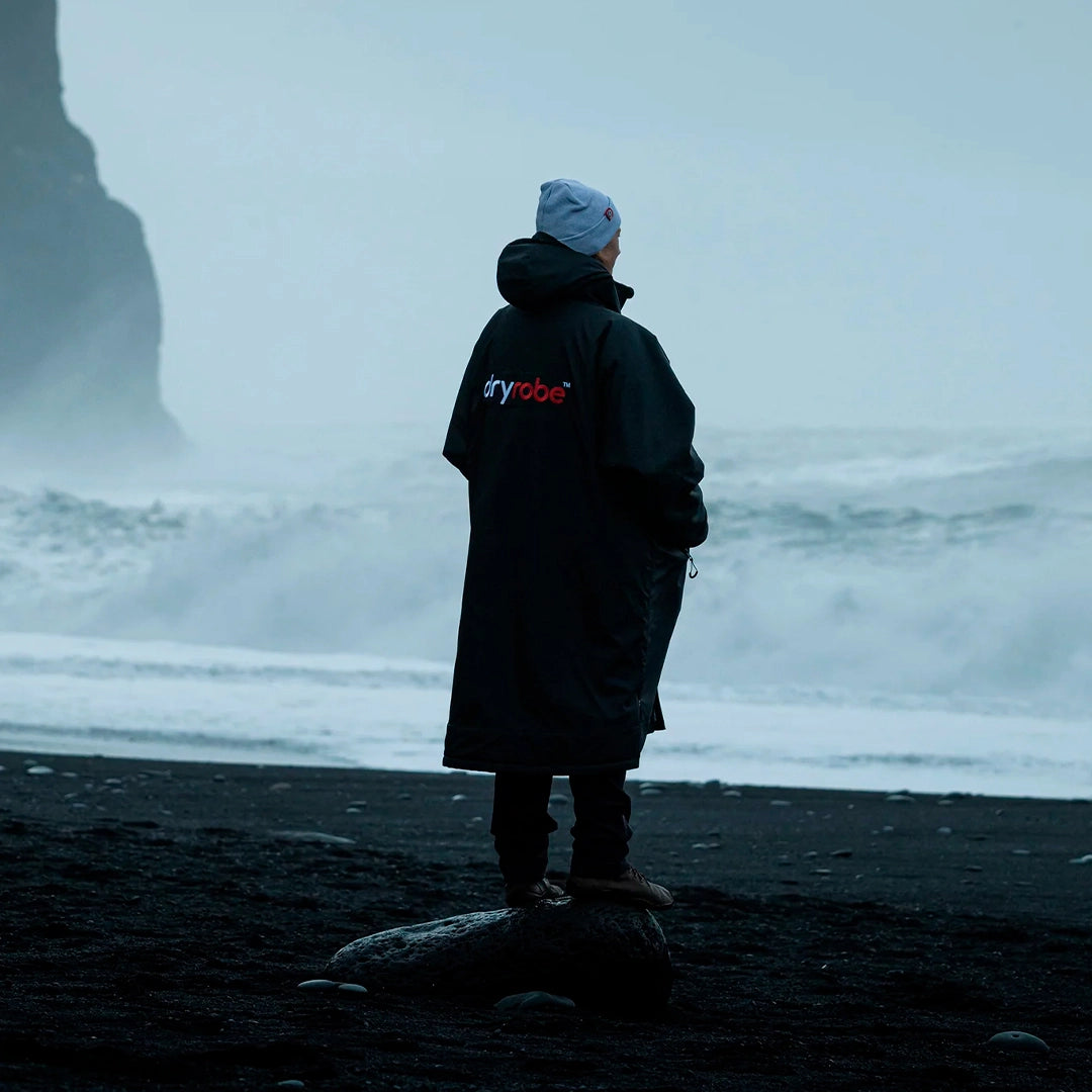Atleta con chaqueta térmica Dryrobe observando el mar, ideal para protegerse del frío después de entrenamientos acuáticos.